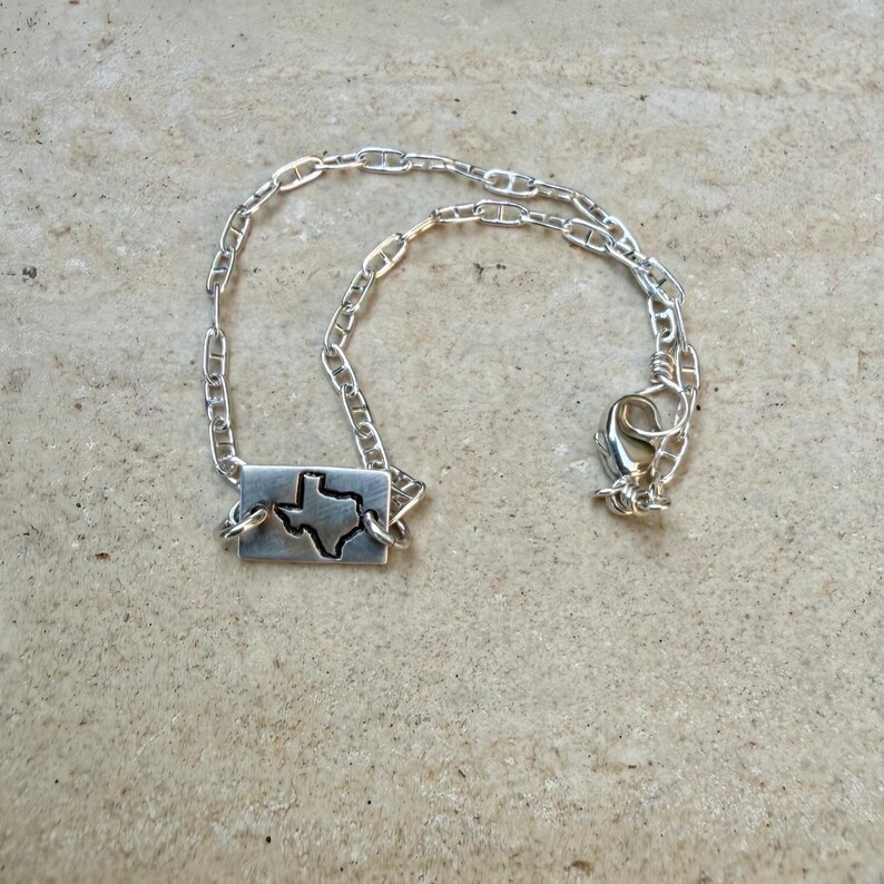 May include: A silver-toned necklace featuring a rectangular pendant with a cutout of the state of Texas. The pendant is connected to a chain-link necklace with a lobster clasp closure. The jewelry is displayed on a neutral-toned surface.