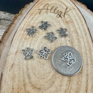 May include: Small, silver-colored butterfly charms arranged on a wooden surface. The charms have an openwork design. A U.S. quarter coin is included for size comparison. The wooden surface has the name "Allyk" written on it.