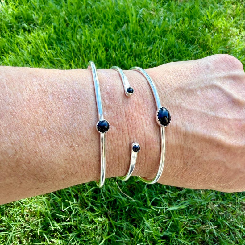 Op de afbeelding: Een set zilverkleurige armbanden met zwarte edelsteenaccenten. De armbanden worden op een pols gedragen tegen een achtergrond van groen gras. De armbanden hebben een minimalistisch ontwerp, met een slanke, gebogen vorm en kleine, ovaalvormige zwarte stenen.