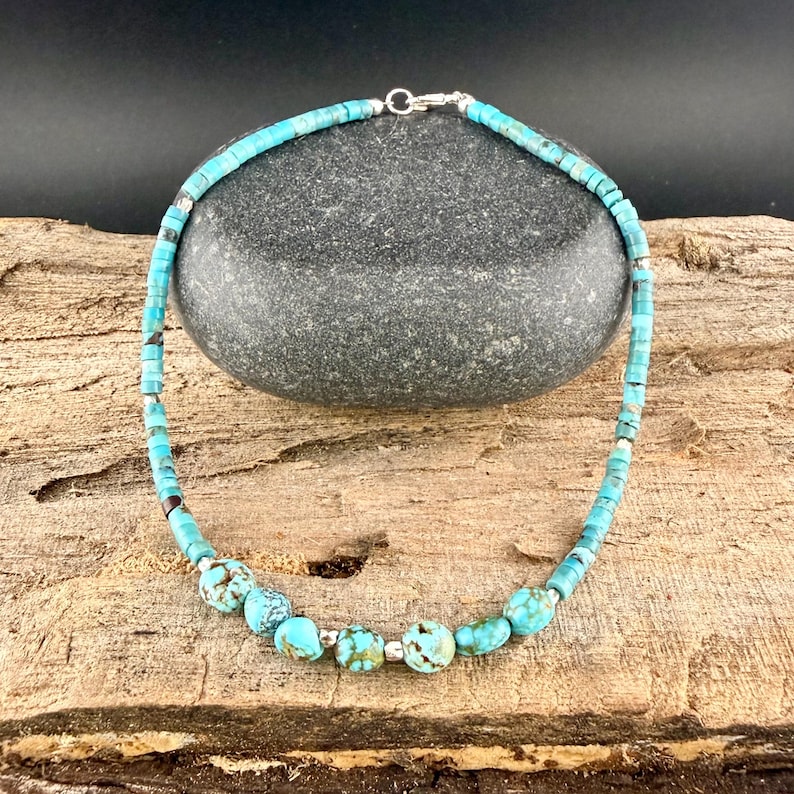 May include: Turquoise beaded necklace with a silver clasp. The necklace features small, square turquoise beads and larger, round turquoise beads with brown and white marbling. The necklace is displayed on a wooden surface.