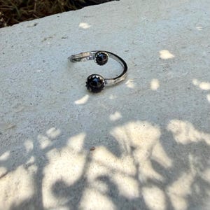 May include: An open-style silver ring featuring two round, dark black stones set in scalloped metal bezels at each end of the band. The silver band has a textured, hammered finish, creating a reflective surface that contrasts with the smooth, dark stones.