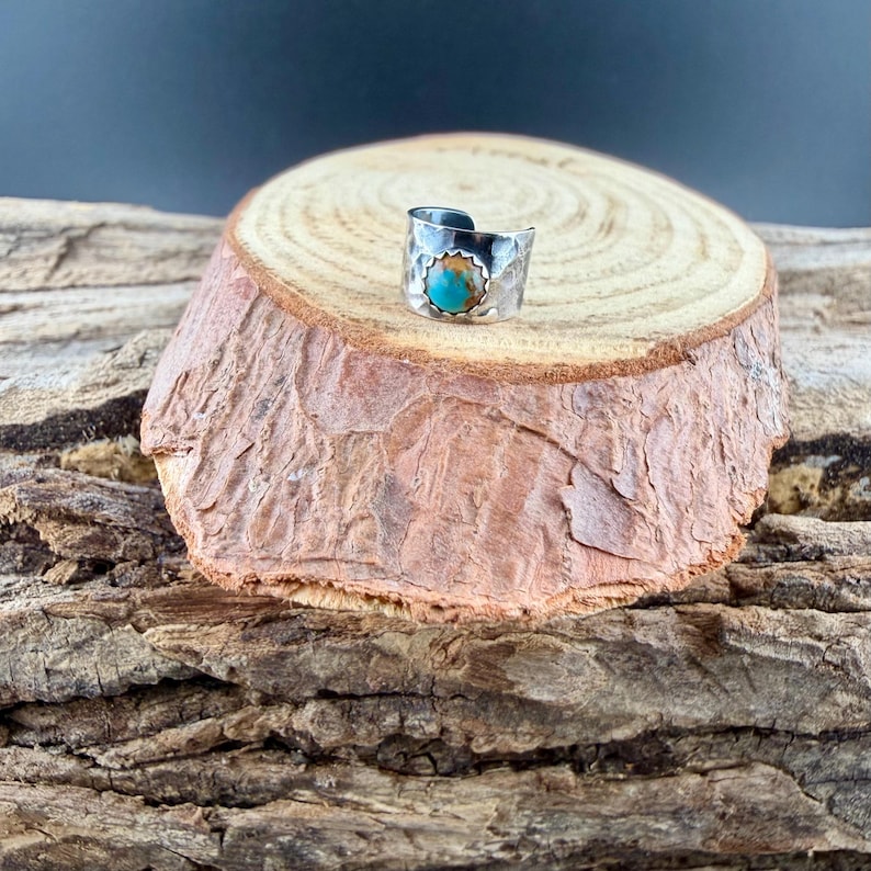 May include: A silver-toned ring with a turquoise stone set in a hammered metal band. The ring is displayed on a small, round, wooden surface with a natural bark edge. The turquoise stone is round and set in a decorative bezel.