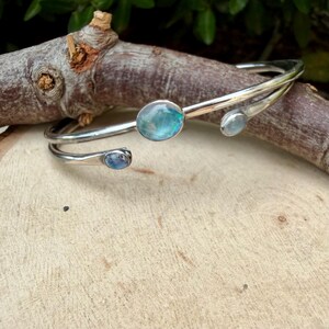 K&ouml;nnte beinhalten: Ein silberfarbenes Armreif-Armband mit drei ovalen, irisierenden blau-gr&uuml;nen Edelsteinen. Das Armband schmiegt sich um das Handgelenk, wobei die Edelsteine entlang des Bandes angeordnet sind. Das Armband wird auf einer Holzoberfl&auml;che pr&auml;sentiert.