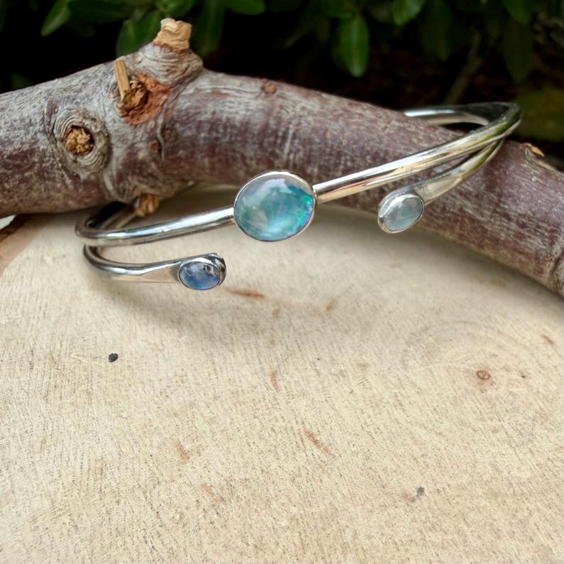 K&ouml;nnte beinhalten: Ein silberfarbenes Armreif-Armband mit drei ovalen, irisierenden blau-gr&uuml;nen Edelsteinen. Das Armband schmiegt sich um das Handgelenk, wobei die Edelsteine entlang des Bandes angeordnet sind. Das Armband wird auf einer Holzoberfl&auml;che pr&auml;sentiert.