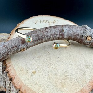 May include: A gold-colored cuff bracelet with turquoise-colored stones. The open-ended bracelet has a curved design with a stone at each end. The bracelet is displayed on a wooden surface with the name "Ally K" etched into the wood.