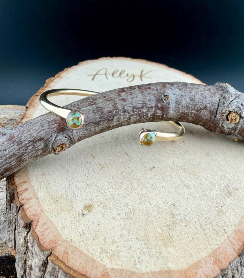 May include: A gold-colored cuff bracelet with turquoise-colored stones. The open-ended bracelet has a curved design with a stone at each end. The bracelet is displayed on a wooden surface with the name "Ally K" etched into the wood.