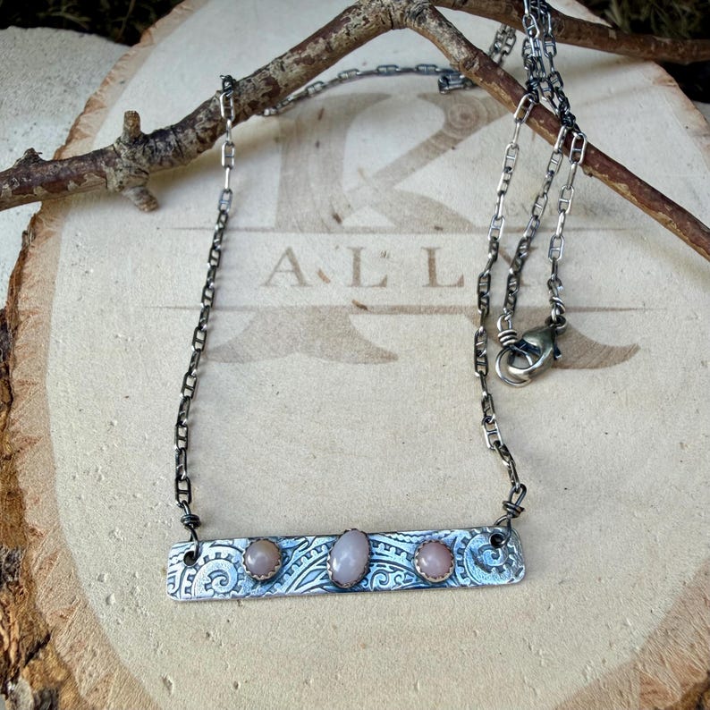 May include: A silver-toned metal bar necklace featuring three small, smooth, pale pink oval stones set in a row. The rectangular bar has an intricate, embossed swirling pattern and is suspended from a dark, oxidized silver-toned chain.
