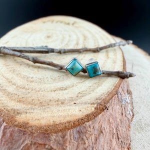 May include: A pair of square turquoise stud earrings with silver settings. The earrings are displayed on a wooden surface with a small twig. The turquoise stones have a mottled appearance with shades of blue and brown.
