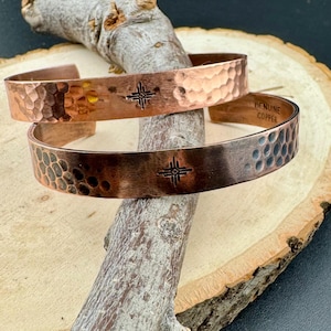 May include: Two copper cuff bracelets with hammered textures and a stamped geometric design. One bracelet has the text "GENUINE COPPER". The bracelets are displayed on a wooden surface.