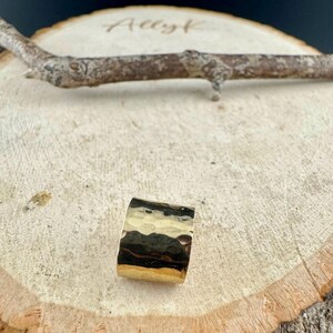 May include: A gold ring with a hammered texture. The ring is rectangular and rests on a wooden surface. The ring's surface reflects light, creating highlights and shadows.