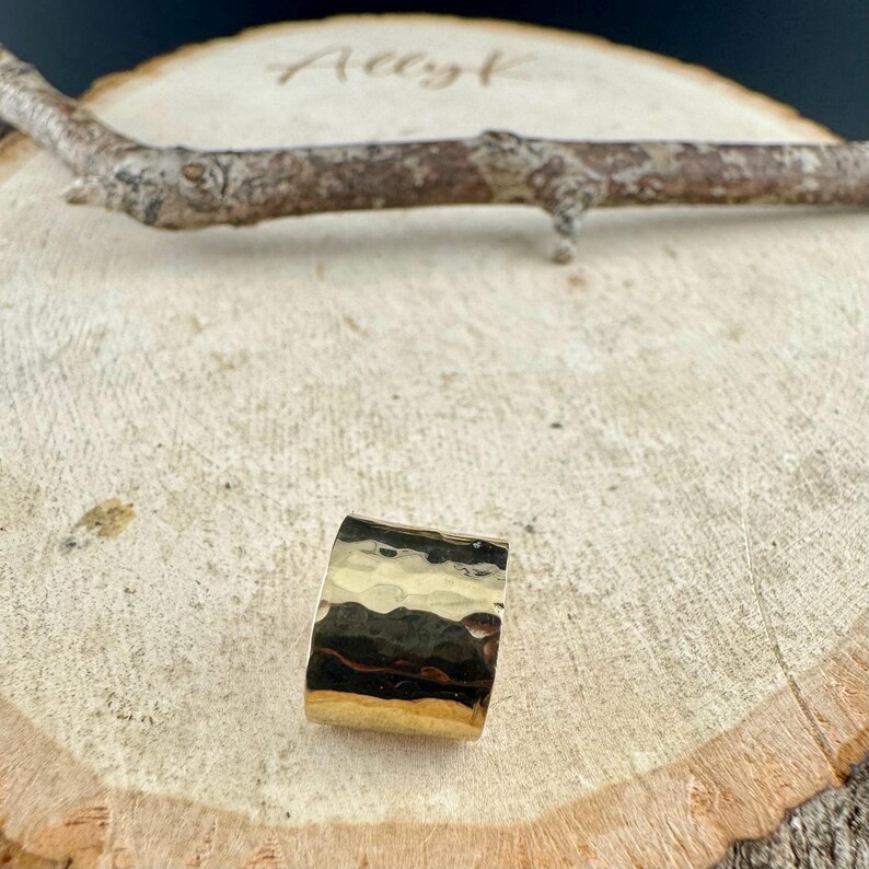 May include: A gold ring with a hammered texture. The ring is rectangular and rests on a wooden surface. The ring's surface reflects light, creating highlights and shadows.