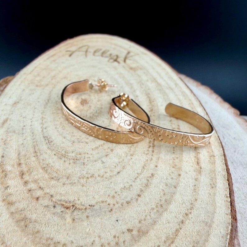 May include: A pair of gold-colored hoop earrings with etched floral and geometric patterns. The earrings are displayed on a wooden surface with a natural grain and the name "Ally K" lightly etched into the wood.