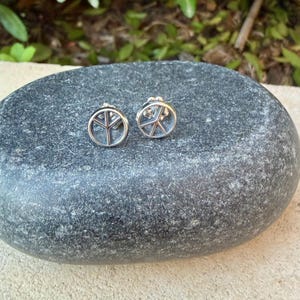 Puede incluir: Un par de pendientes de plata. Cada pendiente presenta un dise&ntilde;o de s&iacute;mbolo de la paz dentro de un marco circular. Los pendientes se muestran sobre una superficie de piedra gris texturizada.