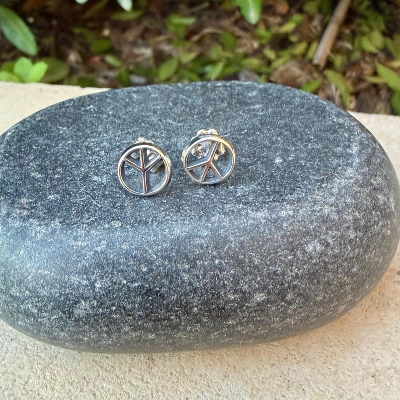 Puede incluir: Un par de pendientes de plata. Cada pendiente presenta un dise&ntilde;o de s&iacute;mbolo de la paz dentro de un marco circular. Los pendientes se muestran sobre una superficie de piedra gris texturizada.