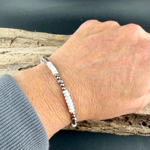 May include: White beaded bracelet with silver-colored accents. The bracelet features small, round white beads interspersed with silver-colored beads and decorative elements. The bracelet is shown on a wrist.