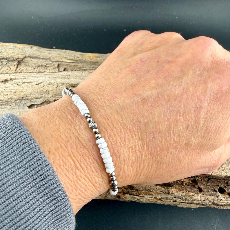 May include: White beaded bracelet with silver-colored accents. The bracelet features small, round white beads interspersed with silver-colored beads and decorative elements. The bracelet is shown on a wrist.