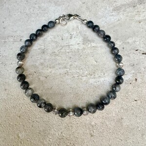 May include: A beaded bracelet featuring round, dark gray, mottled stone beads interspersed with small, ribbed, silver-toned metal spacer beads. The bracelet is secured with a silver-toned metal lobster claw clasp and a circular jump ring.