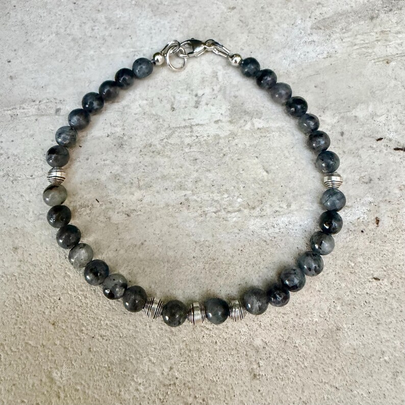 May include: A beaded bracelet featuring round, dark gray, mottled stone beads interspersed with small, ribbed, silver-toned metal spacer beads. The bracelet is secured with a silver-toned metal lobster claw clasp and a circular jump ring.