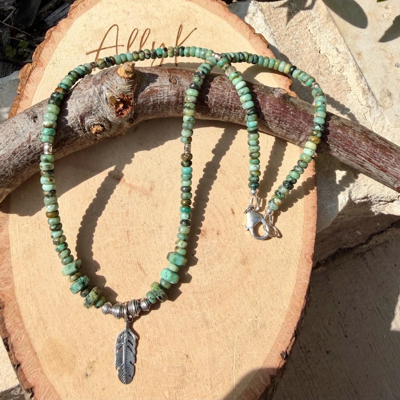May include: A turquoise beaded necklace with a silver feather pendant. The necklace is displayed on a wooden surface. The beads are a mix of green and brown tones. The necklace has a silver clasp.