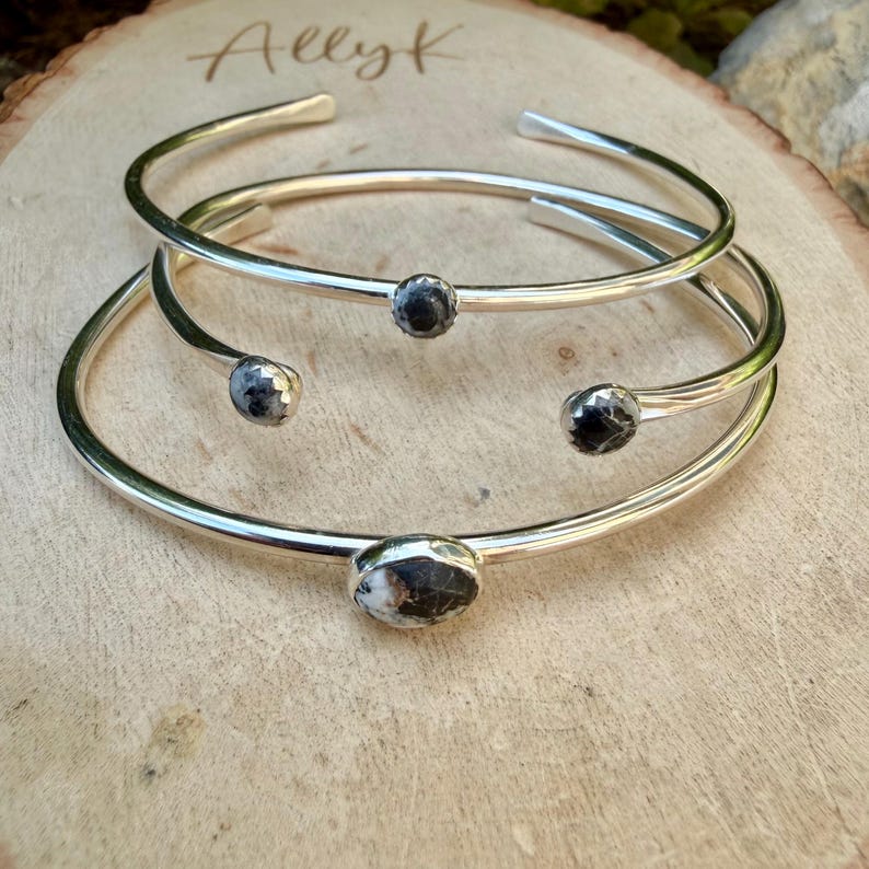 May include: Three silver-toned, open-ended bangles with dark, speckled stone accents. The bangles are stacked on a light-colored wooden surface. The top of the wood has the handwritten text "Ally K".