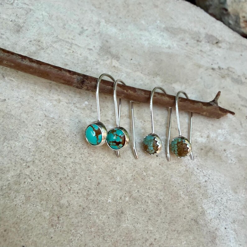 May include: Two pairs of silver drop earrings featuring round turquoise stones with brown matrix patterns. Each earring has a simple silver hook wire design. The stones in the first pair are larger and brighter blue, while the second pair is smaller.