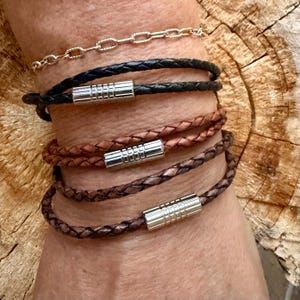 May include: A set of four bracelets consisting of one thin gold-tone chain and three braided leather cords in black, tan, and dark brown. Each leather bracelet features a cylindrical silver-tone metal magnetic clasp with a ribbed texture.