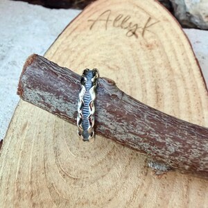 May include: A silver ring with a textured, oxidised finish featuring a repeating vertical grooved pattern along the band. The ring is positioned around a brown, cylindrical wooden branch that has a rough, natural bark surface texture.