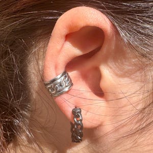 May include: Close-up of an ear adorned with two pieces of jewelry. A silver ear cuff with a chain design sits on the upper ear, and a matching chain-link hoop earring is in the earlobe. The jewelry is made of a shiny, metallic material.