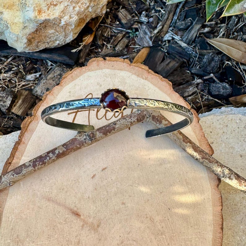Puede incluir: Una pulsera de plata con textura martillada y una gema grande, redonda y de color rojo oscuro. La pulsera es abierta y descansa sobre una superficie de madera, con la palabra "Tuca" visible.