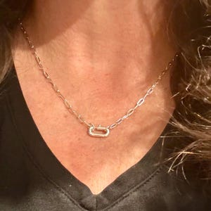 Puede incluir: Un collar de cadena metálica con eslabones ovalados alargados y un cierre rectangular prominente situado en la parte central delantera. El collar tiene un acabado pulido y descansa sobre el cuello, mostrando un diseño sencillo y uniforme.