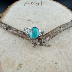 May include: A silver ring with an oval turquoise stone. The ring has a decorative, engraved design on the band. The ring is displayed on a wooden surface with a small branch.