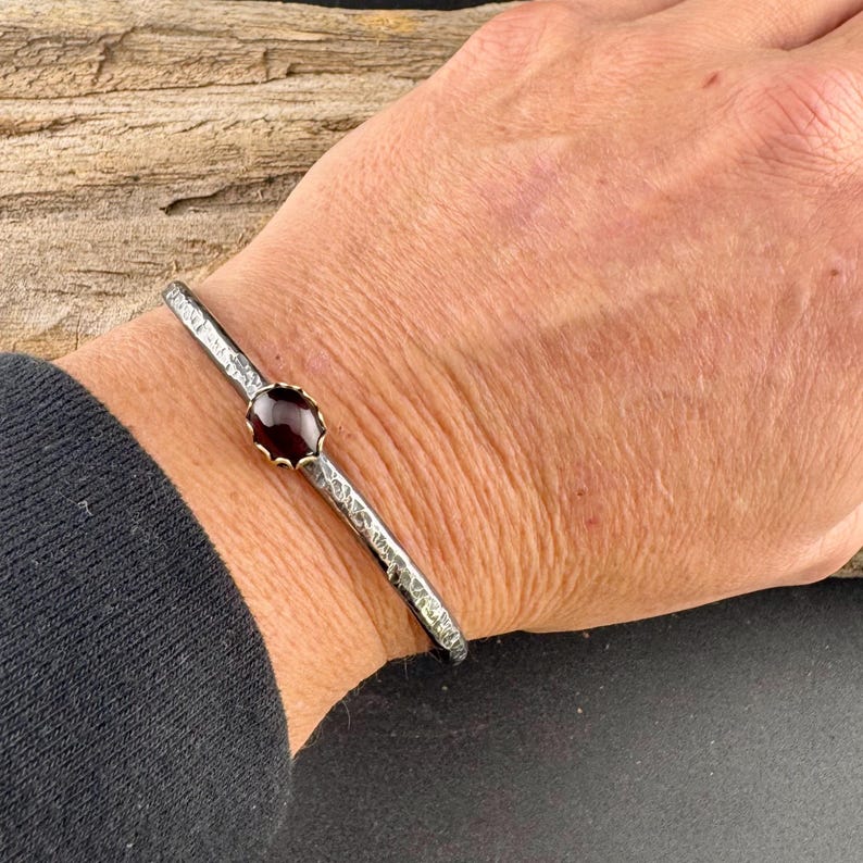 Puede incluir: Un brazalete plateado con textura martillada y una gema central ovalada de color rojo oscuro. El brazalete se lleva en la mu&ntilde;eca, mostrando el dise&ntilde;o y la artesan&iacute;a de la joya.