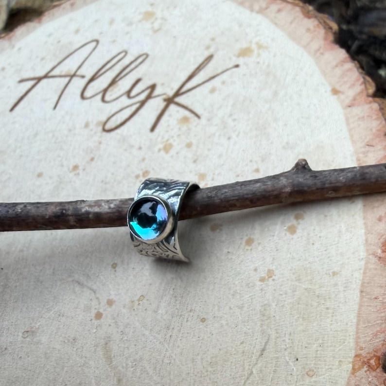 May include: A silver ring with a textured band and a round, iridescent blue-green stone. The ring is displayed on a small wooden branch against a light-colored background. The ring has an engraved floral pattern.