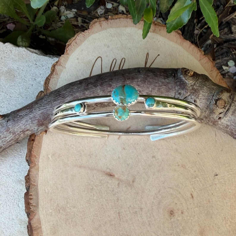 May include: A silver cuff bracelet with three parallel bands. The bracelet features turquoise stones of varying sizes. The bracelet is displayed on a wooden surface with a natural, organic aesthetic.