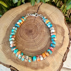 Pode incluir: Uma pulseira de contas com pedras redondas multicoloridas em tons de turquesa, laranja, branco e castanho. As contas s&atilde;o separadas por pequenos espa&ccedil;adores de metal prateado e presas com um fecho lagosta de cor prateada.
