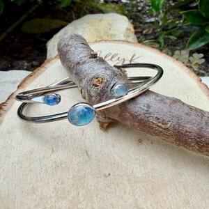 K&ouml;nnte beinhalten: Zwei silberfarbene Metallarmb&auml;nder mit blau und t&uuml;rkis farbenen Steinen. Die Armb&auml;nder sind offen, mit einem geschwungenen Design. Die Steine haben ein durchscheinendes, marmoriertes Aussehen. Die Armb&auml;nder liegen auf einer Holzoberfl&auml;che.