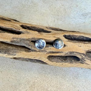 May include: A pair of circular silver stud earrings featuring a stamped diamond pattern in the center, surrounded by small etched lines. The earrings have a polished metallic finish and are displayed on a piece of textured driftwood.