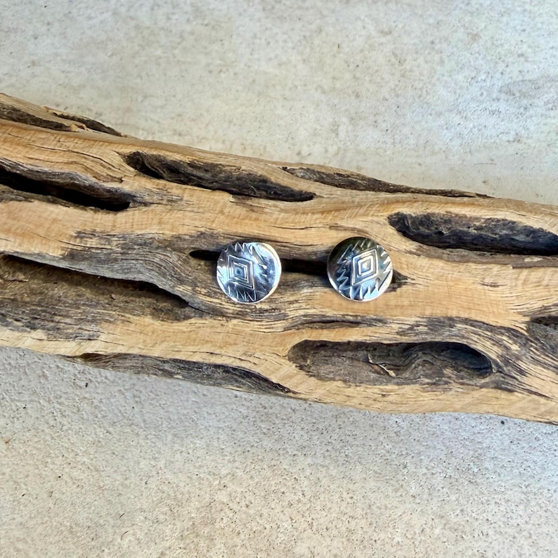 May include: A pair of circular silver stud earrings featuring a stamped diamond pattern in the center, surrounded by small etched lines. The earrings have a polished metallic finish and are displayed on a piece of textured driftwood.