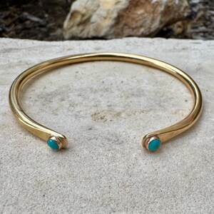 May include: A gold-colored cuff bracelet with turquoise accents. The open-ended bracelet has rounded terminals, each featuring a small, oval turquoise stone. The bracelet is displayed on a light-colored, textured surface.
