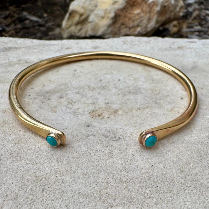 May include: A gold-colored cuff bracelet with turquoise accents. The open-ended bracelet has rounded terminals, each featuring a small, oval turquoise stone. The bracelet is displayed on a light-colored, textured surface.