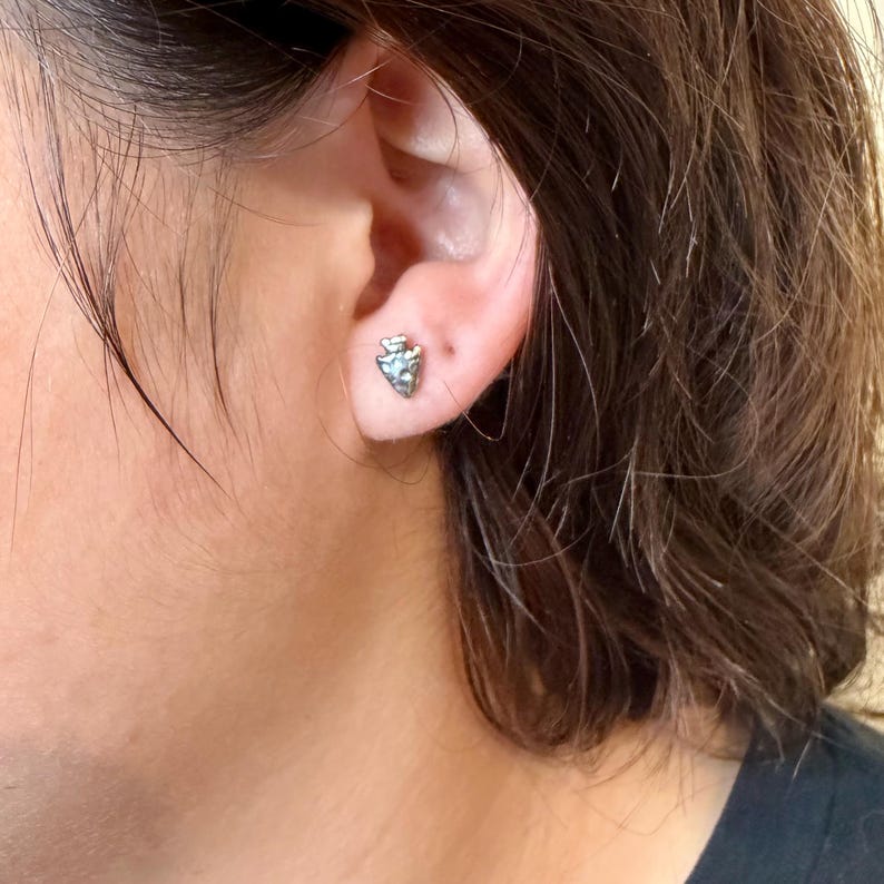 May include: A close-up of a silver arrowhead stud earring. The earring is worn on the ear. The arrowhead design has a textured surface and a small, light-colored stone in the center. The person's skin and hair are visible in the background.