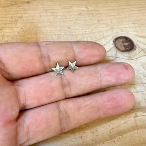May include: A pair of small, five-pointed star stud earrings crafted from a polished, reflective silver-toned metal. Each star features clean, sharp edges and a smooth, metallic surface that catches the light, creating a bright, uniform appearance.