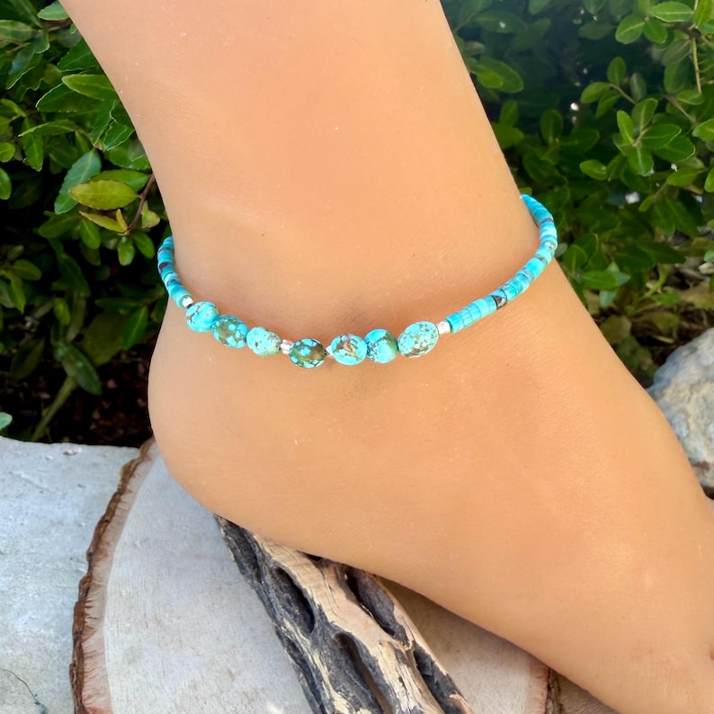 May include: Turquoise beaded anklet with silver accents. The anklet features a mix of small, round turquoise beads and larger, textured turquoise beads. The anklet is worn on an ankle, showcasing the jewelry's design and color.