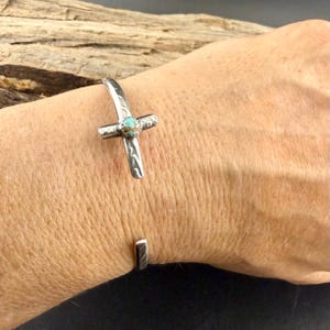 May include: Silver-toned cuff bracelet featuring a cross design with a turquoise stone at the center. The cross has etched details and is the focal point of the open-ended bracelet. The bracelet is shown on a wrist.