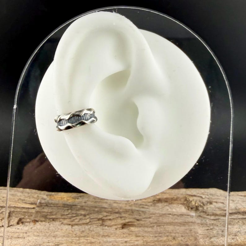 May include: A silver-toned ear cuff with a textured, wave-like design. The cuff is worn on a white ear model, showcasing its intricate details and craftsmanship. The jewelry piece is designed to be worn on the ear.