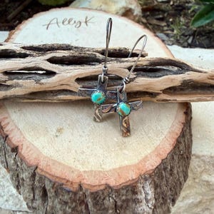 May include: A pair of silver cross earrings with turquoise-colored stones at the center. The earrings are displayed on a wooden surface with the handwritten text "Allyk". The crosses have a textured design and are suspended from curved ear wires.