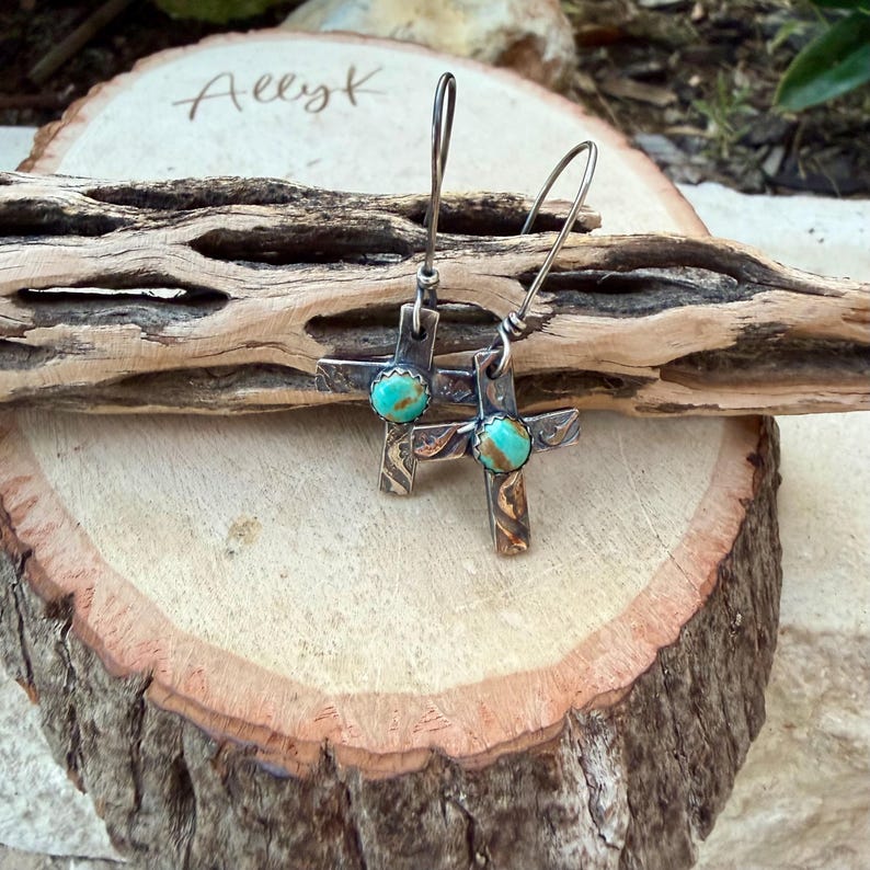 May include: A pair of silver cross earrings with turquoise-colored stones at the center. The earrings are displayed on a wooden surface with the handwritten text "Allyk". The crosses have a textured design and are suspended from curved ear wires.