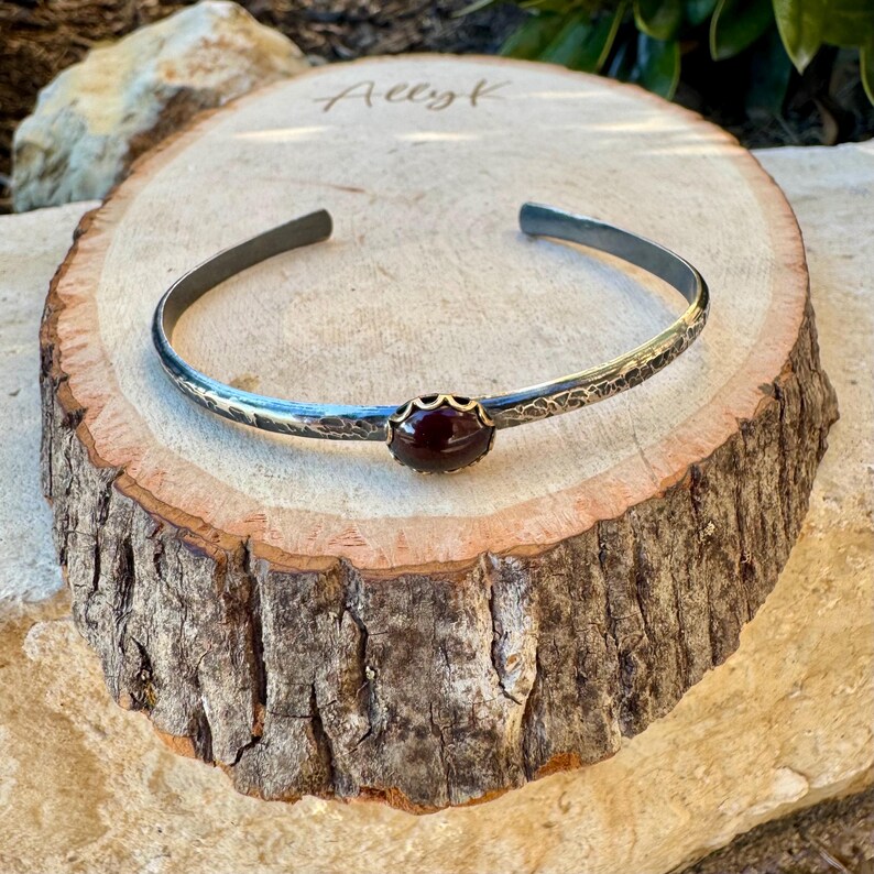 Puede incluir: Una pulsera de plata con una banda texturizada y una piedra preciosa de color rojo oscuro engastada en un bisel dorado. La pulsera se exhibe sobre una rodaja de madera con el nombre "Allyk" grabado.