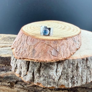 K&ouml;nnte beinhalten: Ein silberner Ring mit einem gro&szlig;en, runden, hellblauen Stein. Der Ring hat ein breites Band mit ge&auml;tzten Details. Der Ring wird auf einer rustikalen Holzoberfl&auml;che mit dunklem Hintergrund pr&auml;sentiert.