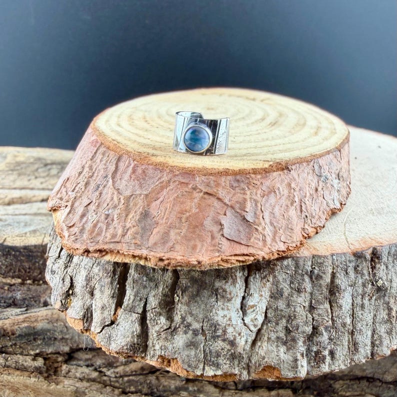 K&ouml;nnte beinhalten: Ein silberner Ring mit einem gro&szlig;en, runden, hellblauen Stein. Der Ring hat ein breites Band mit ge&auml;tzten Details. Der Ring wird auf einer rustikalen Holzoberfl&auml;che mit dunklem Hintergrund pr&auml;sentiert.